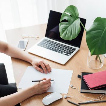 hand hokding a pen with a laptop, notebook, and plant in the background