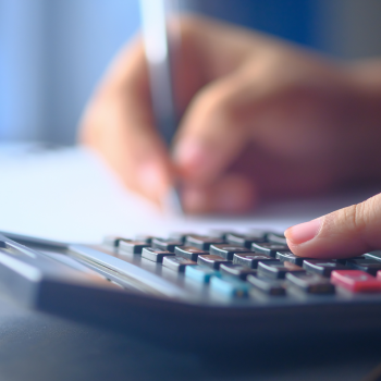 A close up if calculator with a unfocused hand holding a pen in the background.
